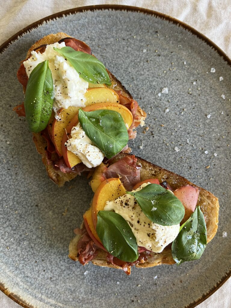 Served Seasonal Nectarine and Buratta Summer Toast. Sourdough toast topped with fig preserves, prosciutto, sliced nectarines, buratta, basil, olive oil, salt and pepper.